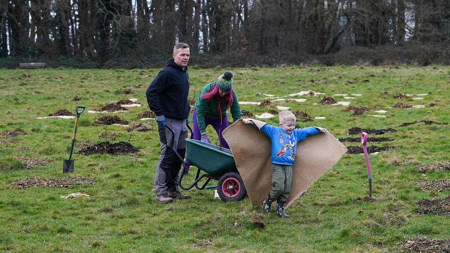 Family in field using spades and wheelbarrow to help plant trees.